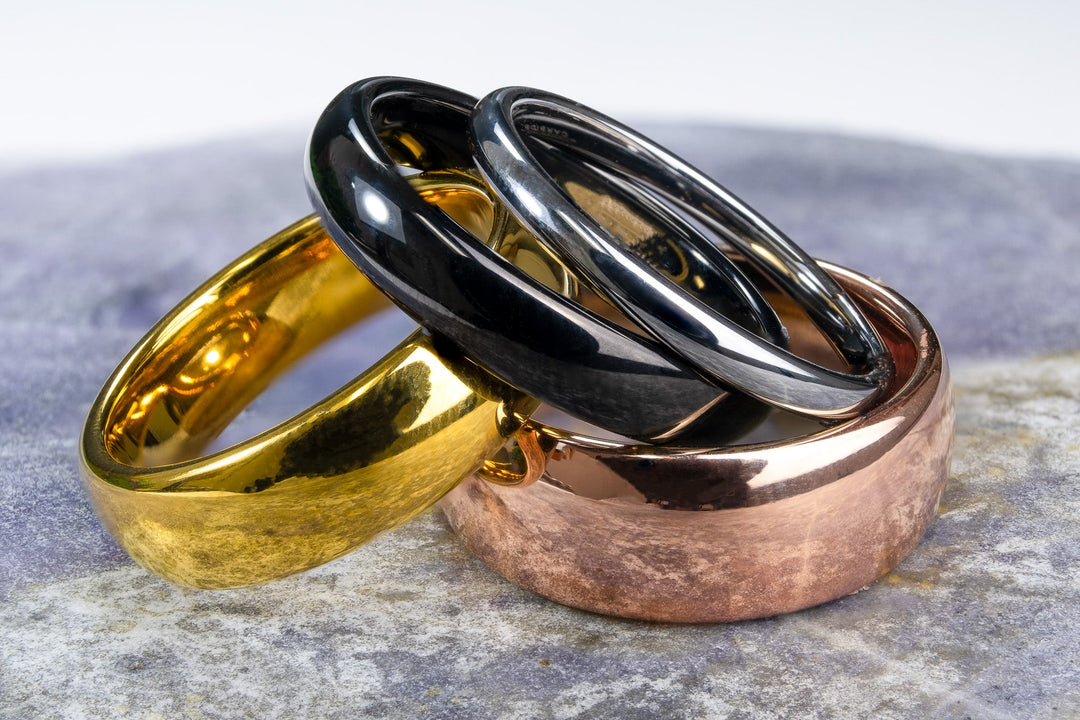 Tungsten wedding bands in polished gold, black, silver and rose finishes displayed on a smooth stone surface.