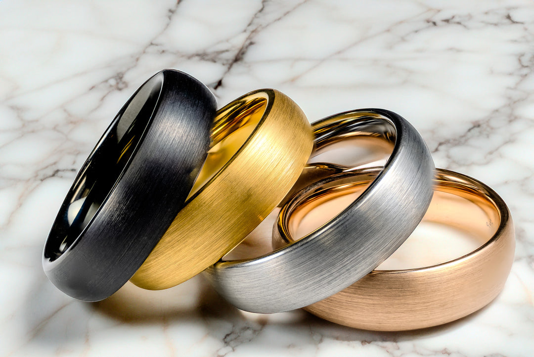 Brushed tungsten wedding bands in black, gold, silver, and rose tones displayed on a marble background.