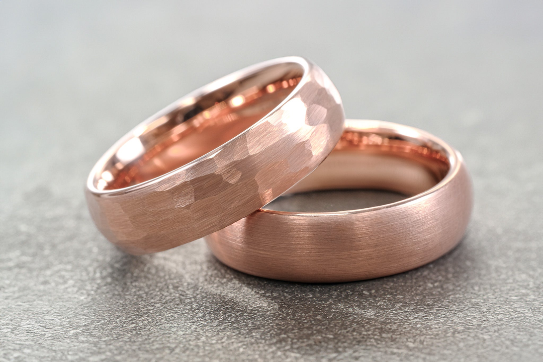 A pair of 6mm rose gold tungsten carbide wedding bands in brushed and hammered finish, laying on top of a grey stone.