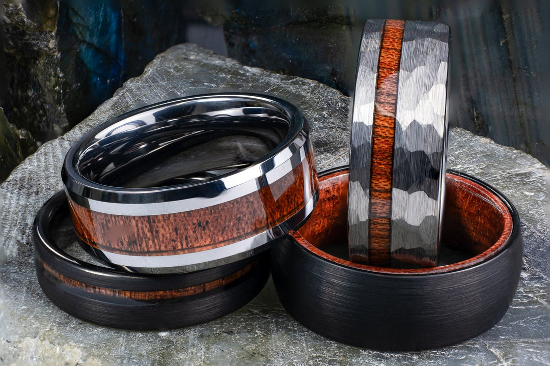 Tungsten wedding bands with authentic Hawaiian Koa wood inlays in black, silver, and polished finishes, displayed on a stone surface.