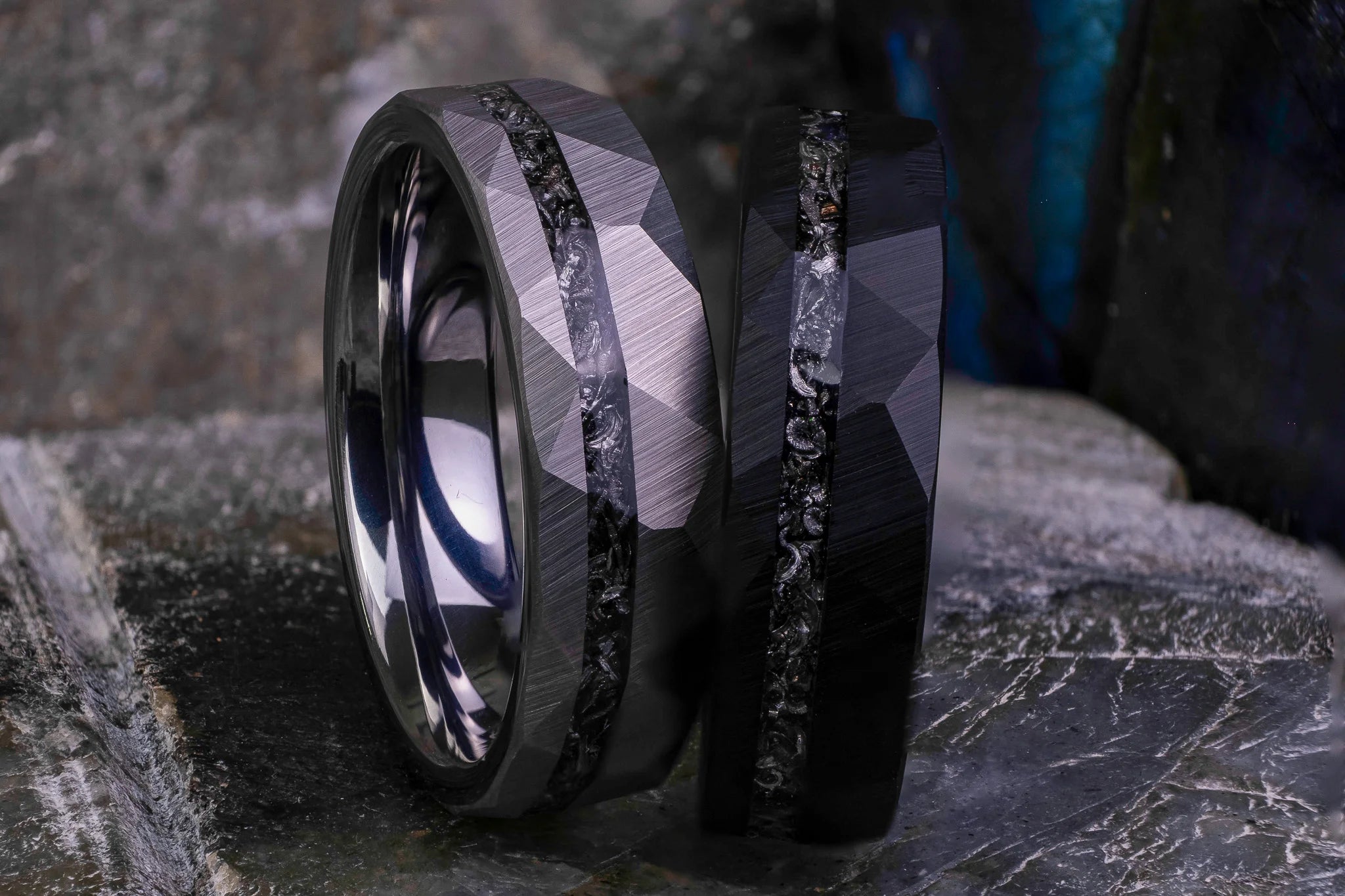 Close-up of The Nova Rift tungsten wedding rings in 8mm hammered black and silver featuring a crushed meteorite stripe and faceted finish, placed on a dark textured surface.