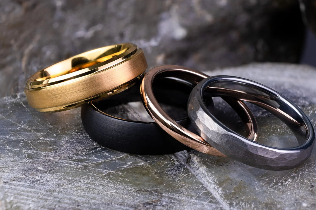  Tungsten carbide wedding bands under $100, featuring brushed gold, matte black, polished rose gold, and hammered silver finishes, arranged on a stone surface.