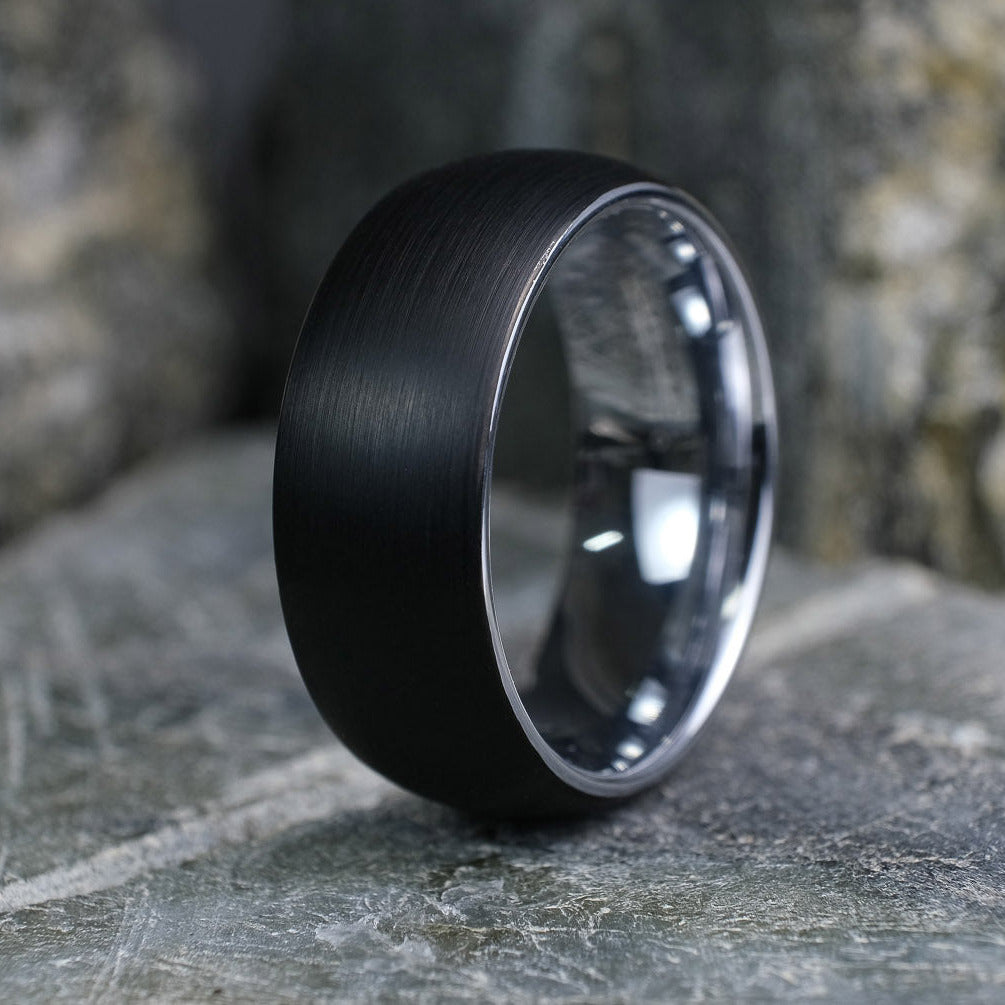Black Brushed tungsten carbide wedding band with a silver polished interior standing up on a textured grey stone.