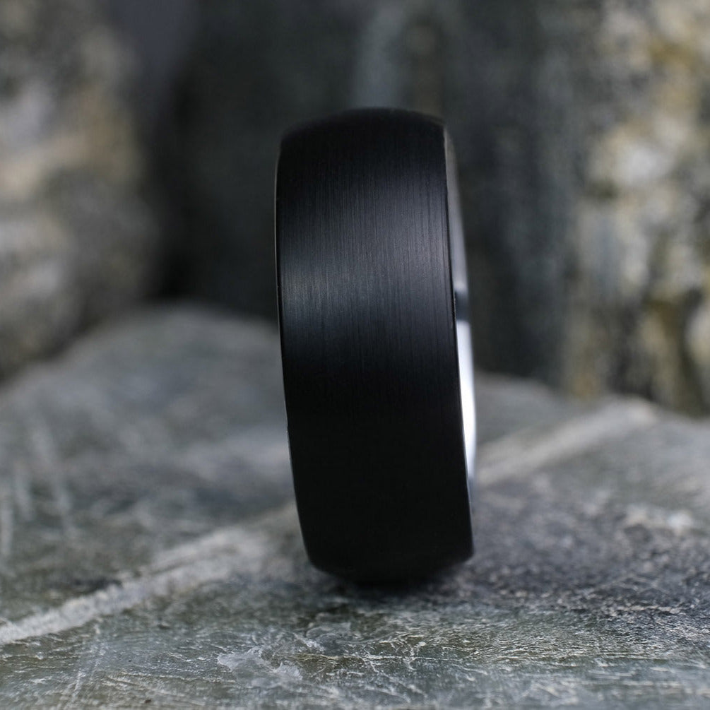 Black Brushed tungsten carbide wedding band with a silver polished interior standing up on a textured grey stone.