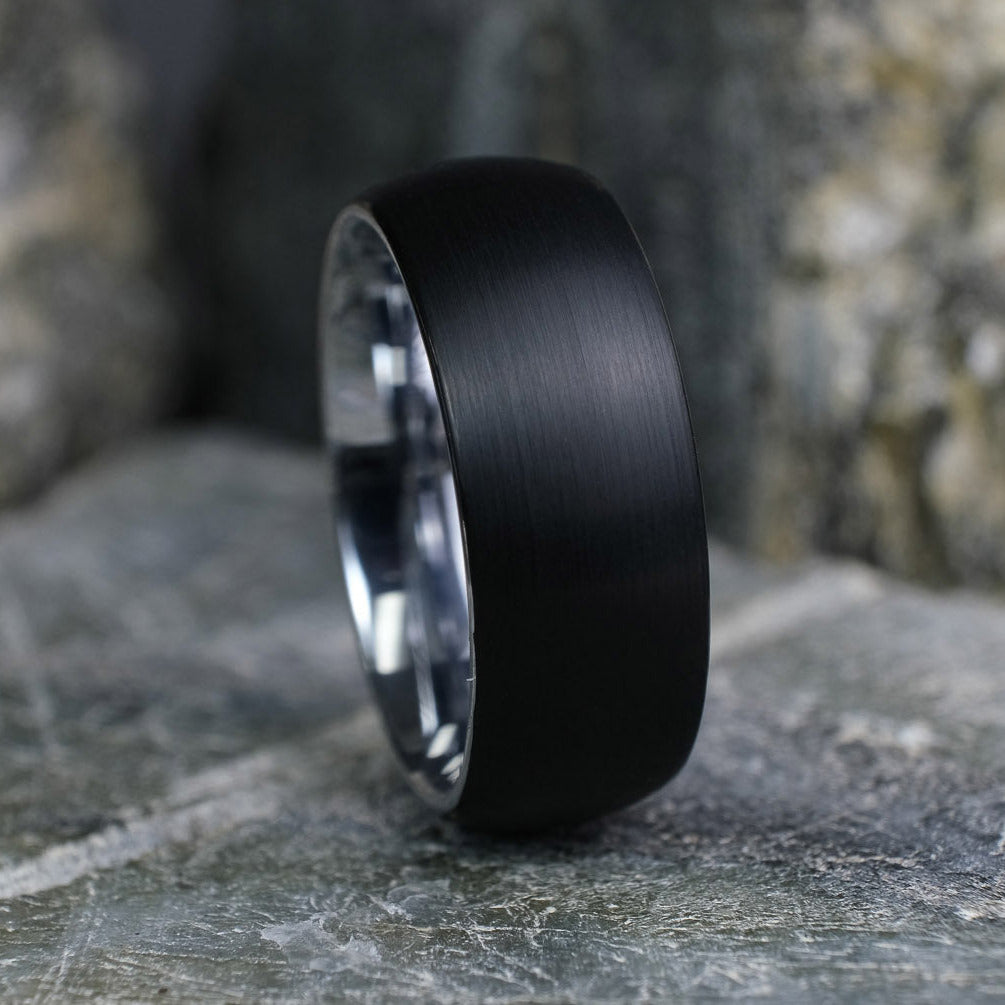 Black Brushed tungsten carbide wedding band with a silver polished interior standing up on a textured grey stone.