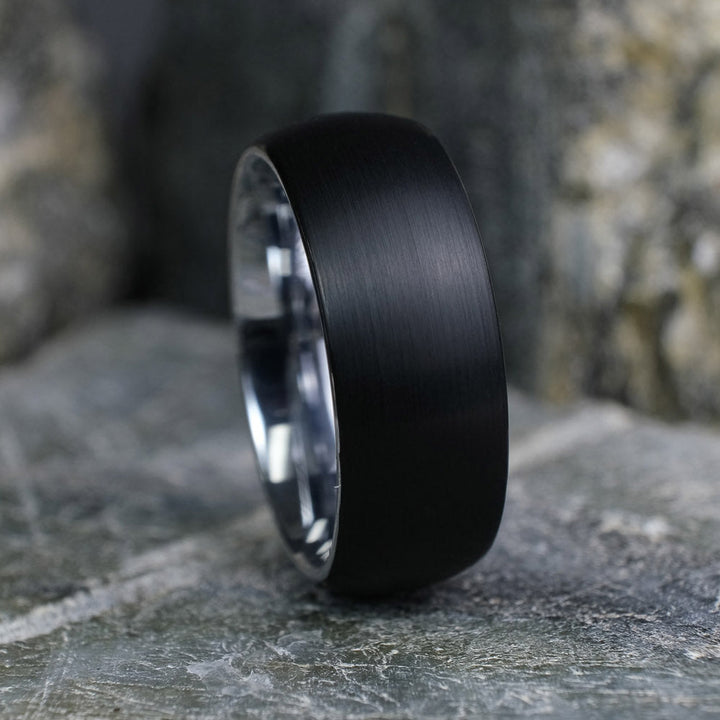 Black Brushed tungsten carbide wedding band with a silver polished interior standing up on a textured grey stone.