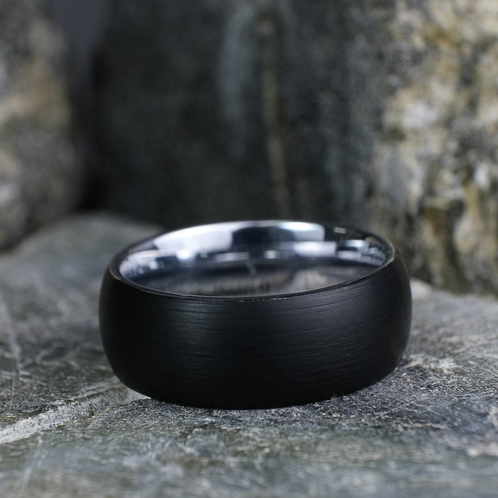 Black Brushed tungsten carbide wedding band with a silver polished interior standing up on a textured grey stone.