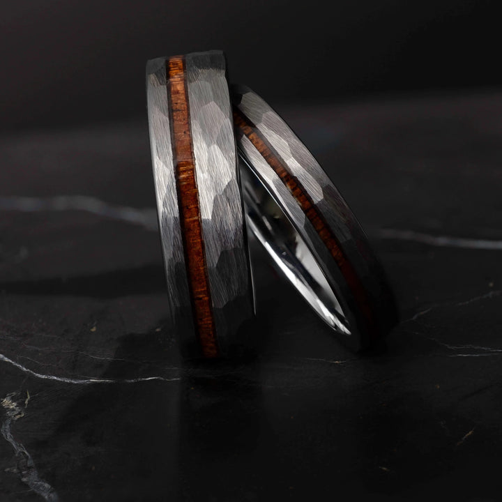Koa wood his and hers silver hammered tungsten carbide wedding bands in 6mm and 4mm on a dark marble slab.