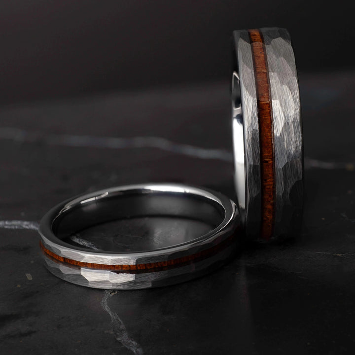 Koa wood his and hers silver hammered tungsten carbide wedding bands in 6mm and 4mm on a dark marble slab.