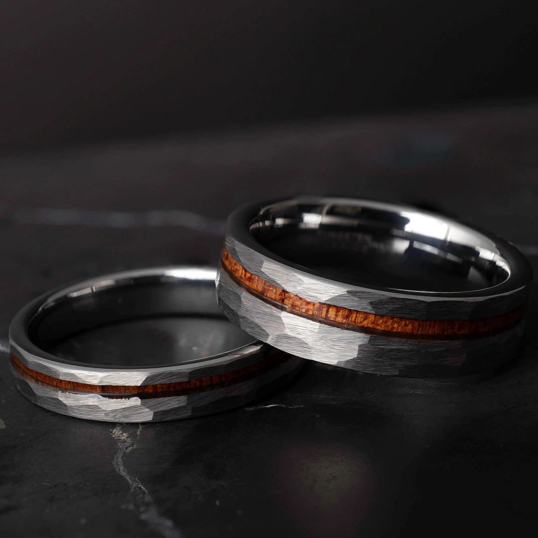 Koa wood his and hers silver hammered tungsten carbide wedding bands in 6mm and 4mm on a dark marble slab.