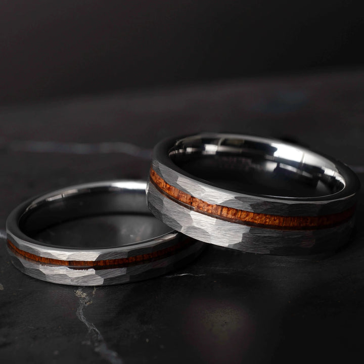 Koa wood his and hers silver hammered tungsten carbide wedding bands in 6mm and 4mm on a dark marble slab.